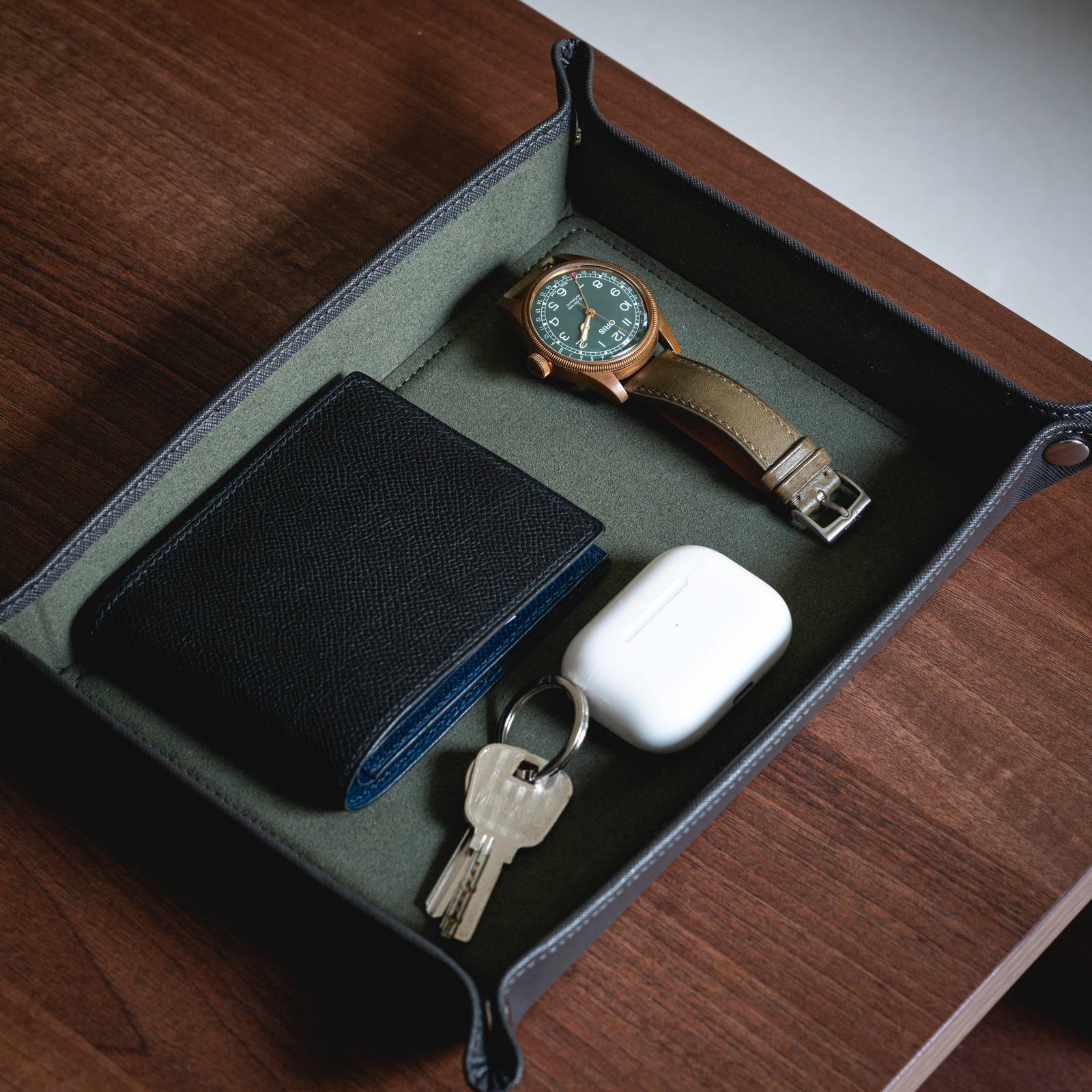Delugs valet tray in grey with dark green interior holding a bronze watch, wallet, keys and earbuds